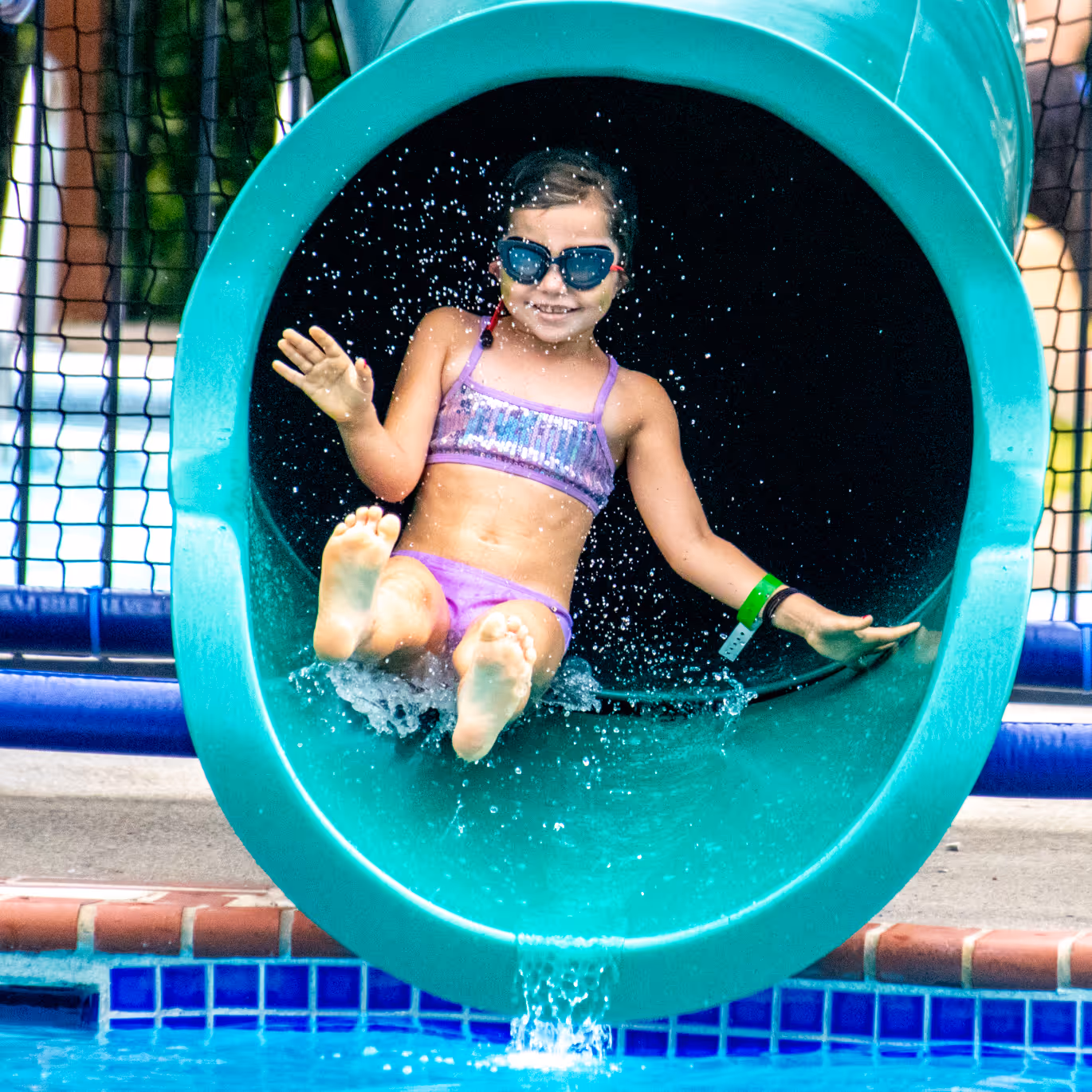 Child wearing a purple swimsuit and sunglasses exiting a green water slide into a pool with water splashing.