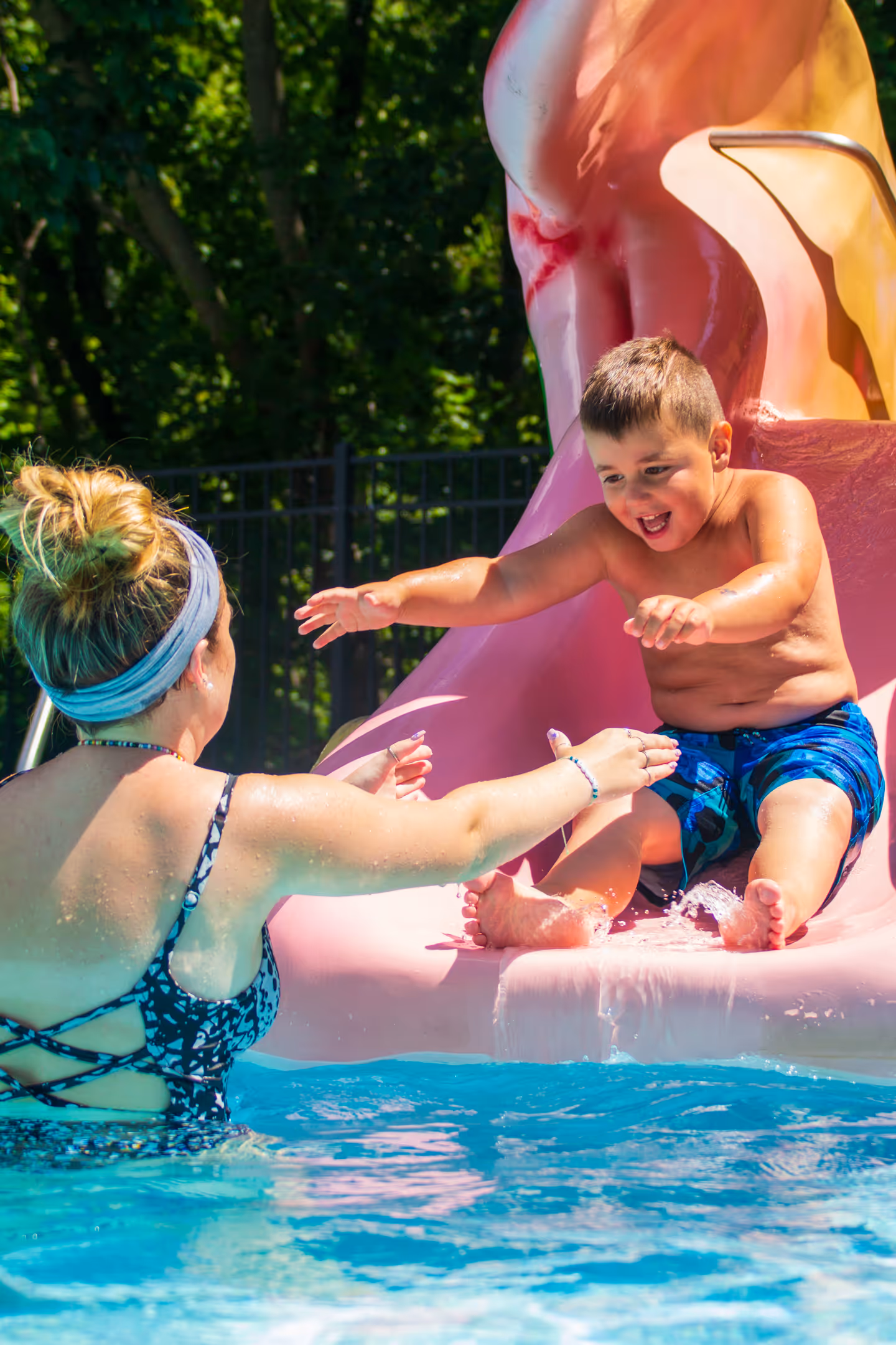 Young boy in blue swim trunks sliding down a pink water slide into a pool, reaching out to a woman in a swimsuit waiting with open arms.