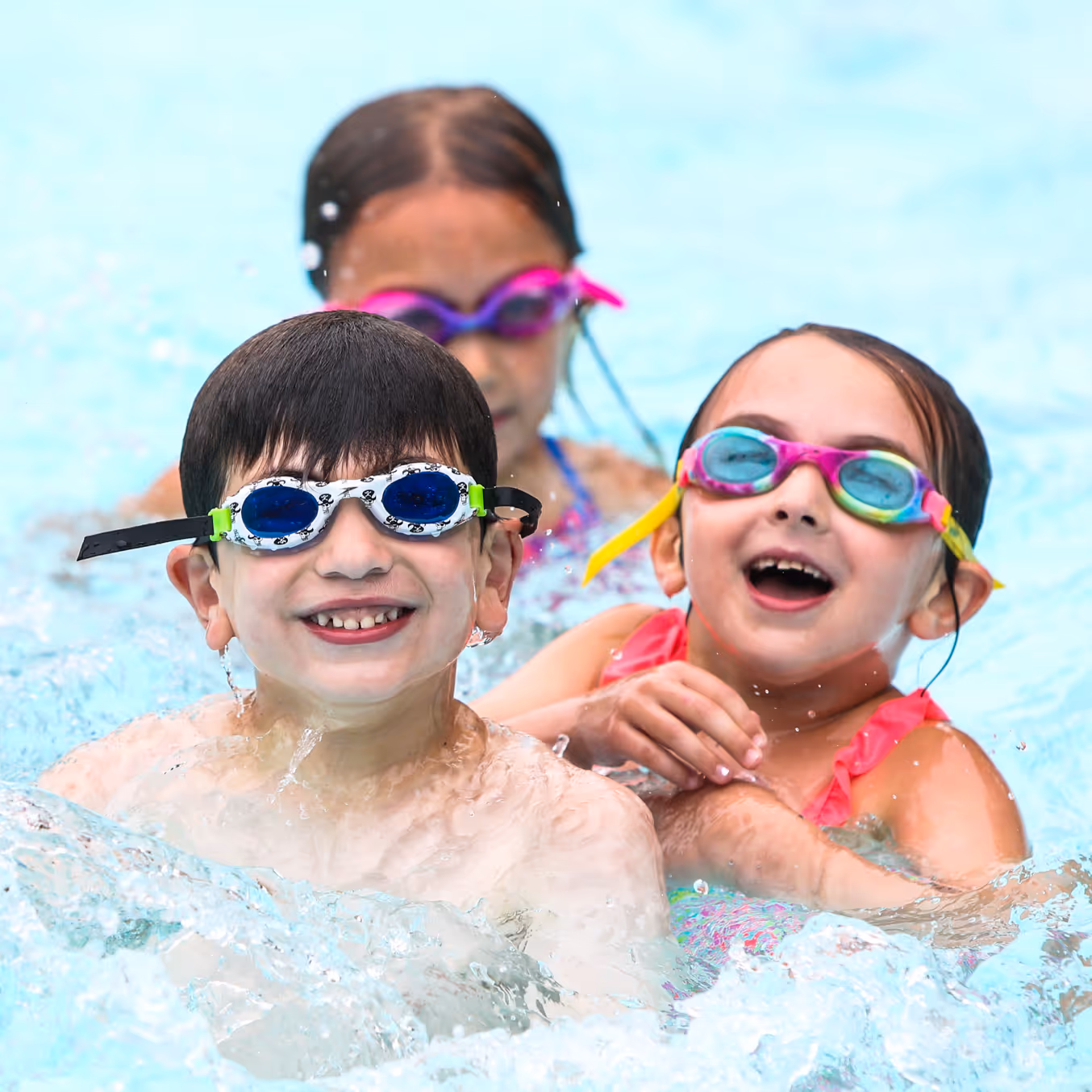 Three children wearing colorful swim goggles smiling and playing in a swimming pool.