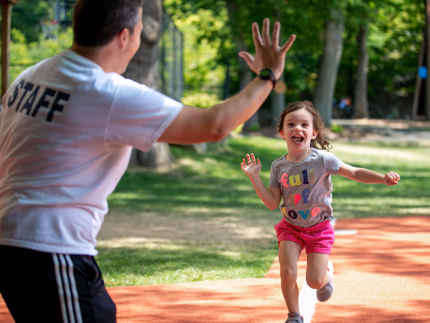 Smiling young girl running towards a man in a white staff T-shirt for a high five on an outdoor path.