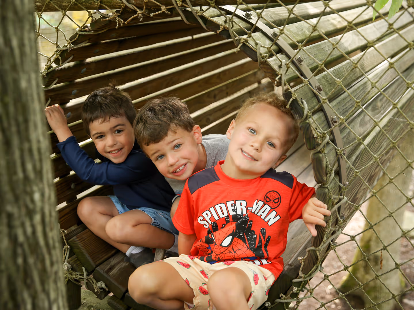 Three young boys smiling inside a wooden tunnel structure at a playground.