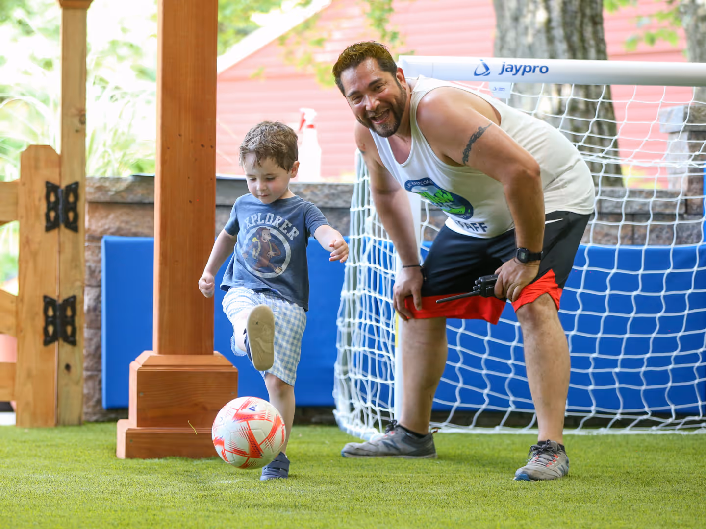 Young boy kicking a soccer ball indoors while a smiling man watches near a small goal net.