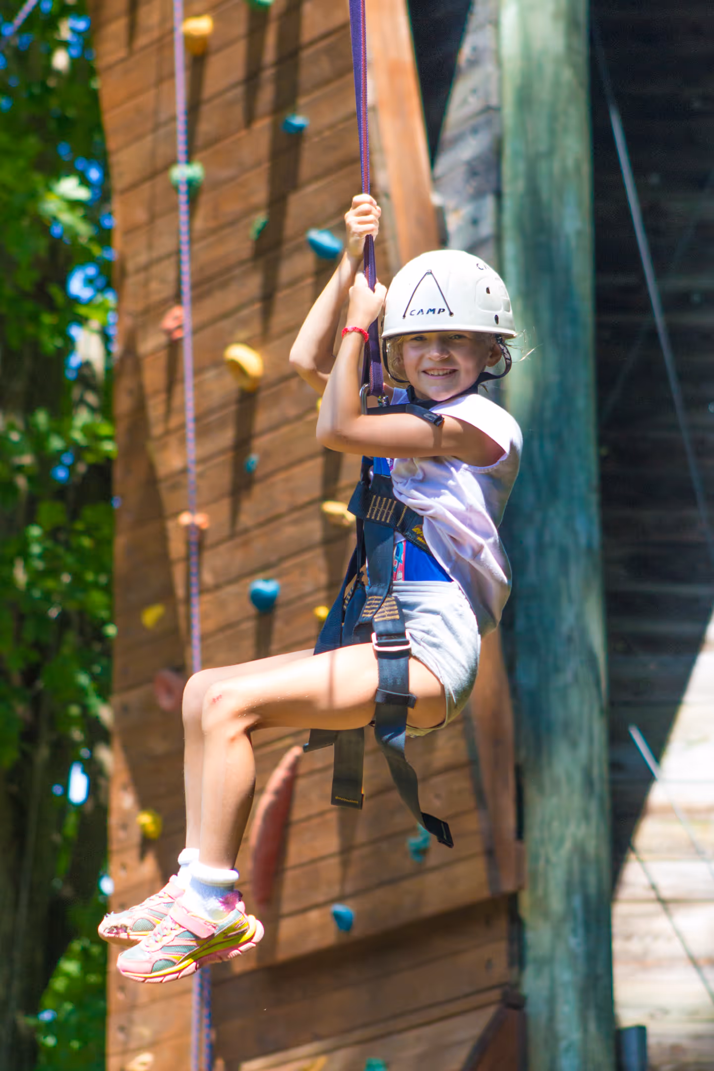 Smiling girl wearing a helmet and safety harness ziplining outdoors near a climbing wall.
