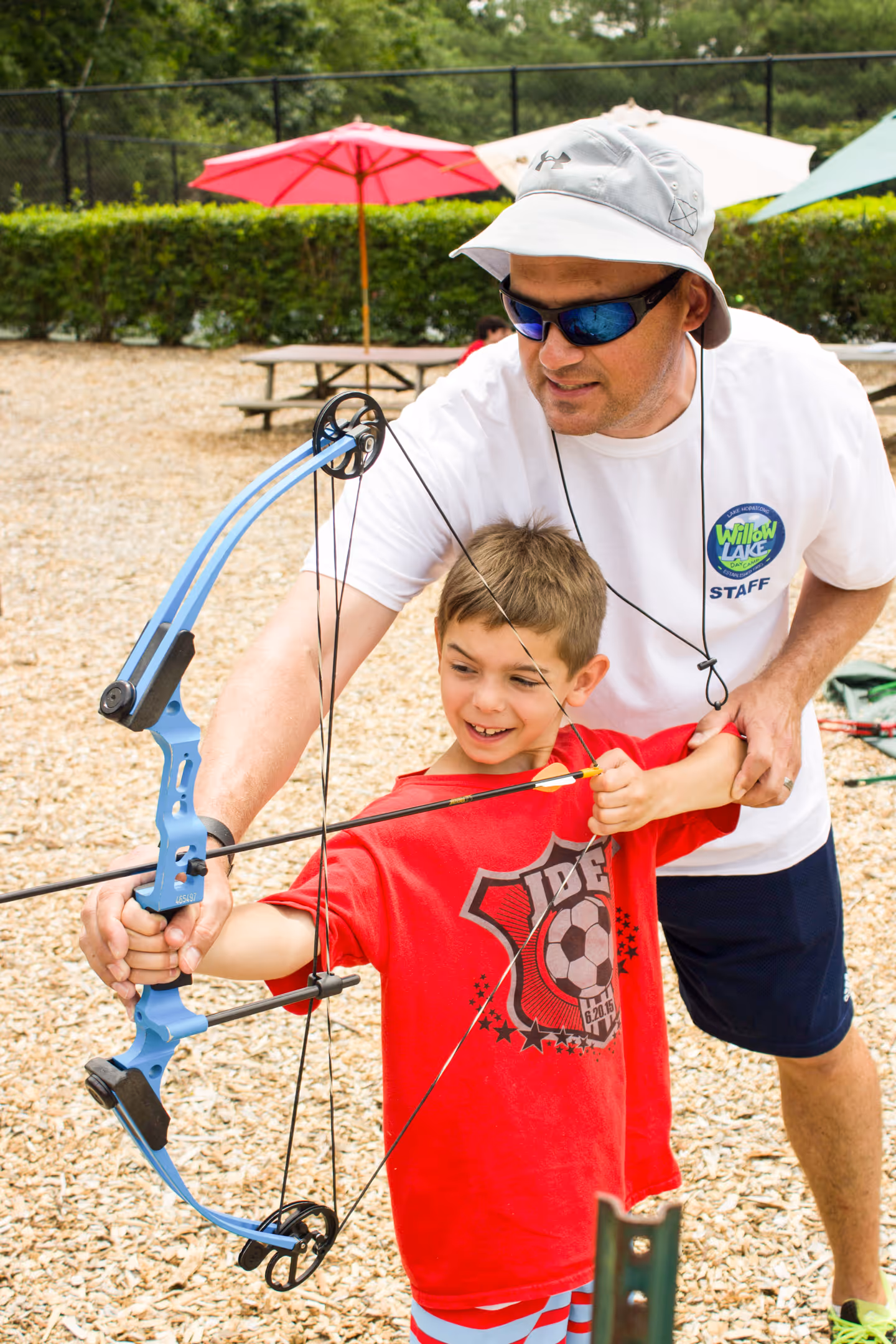 Camp staff member wearing sunglasses and a hat helps a smiling boy in a red shirt aim a blue compound bow for archery outside.