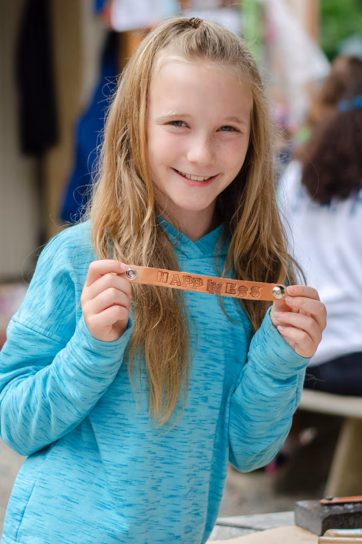 Smiling girl with long blonde hair wearing a blue hoodie holding a leather bracelet engraved with the word 'HAPPINESS'.