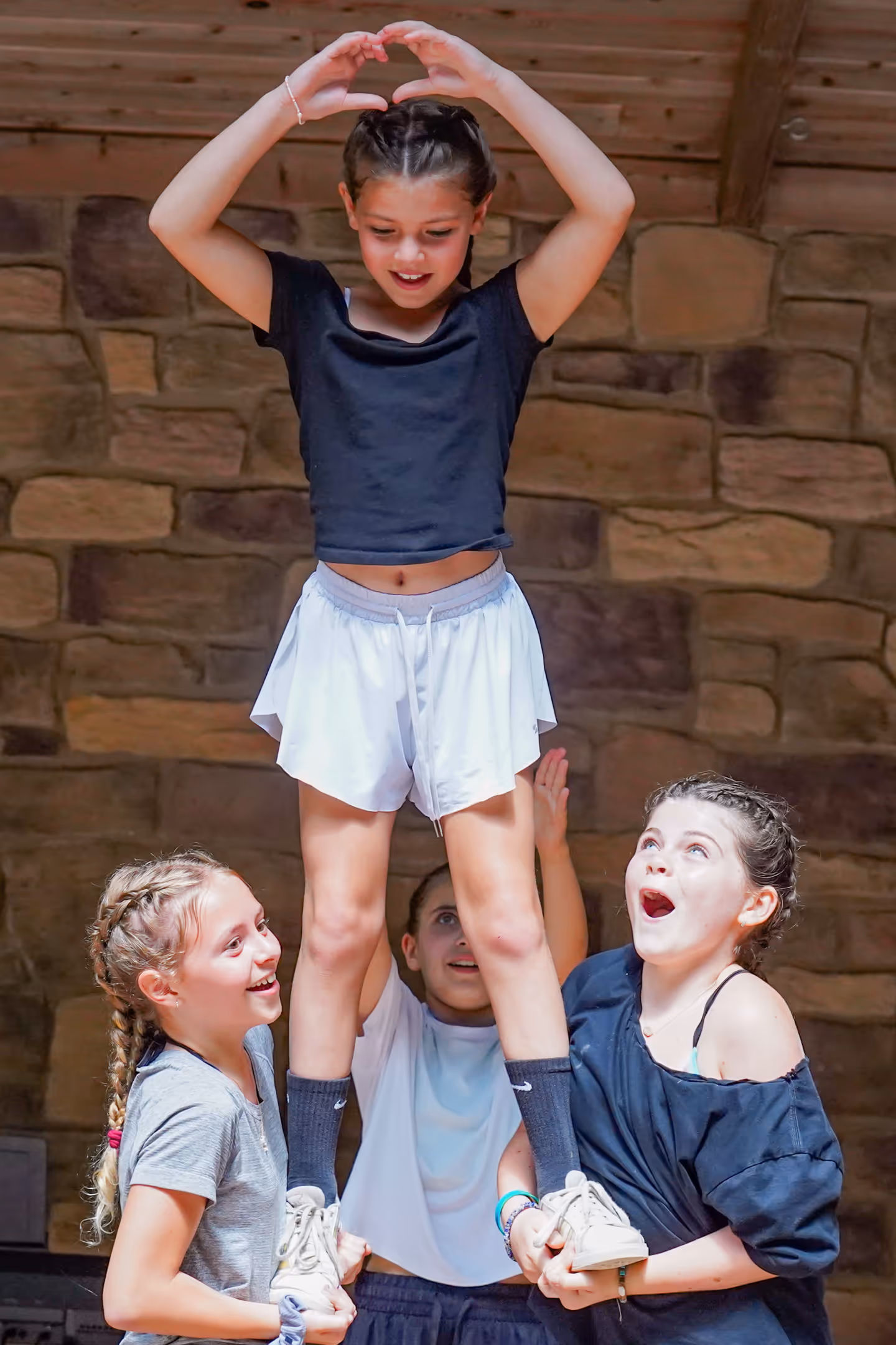 Three girls lifting another girl in a cheerleading stunt with a stone wall background.