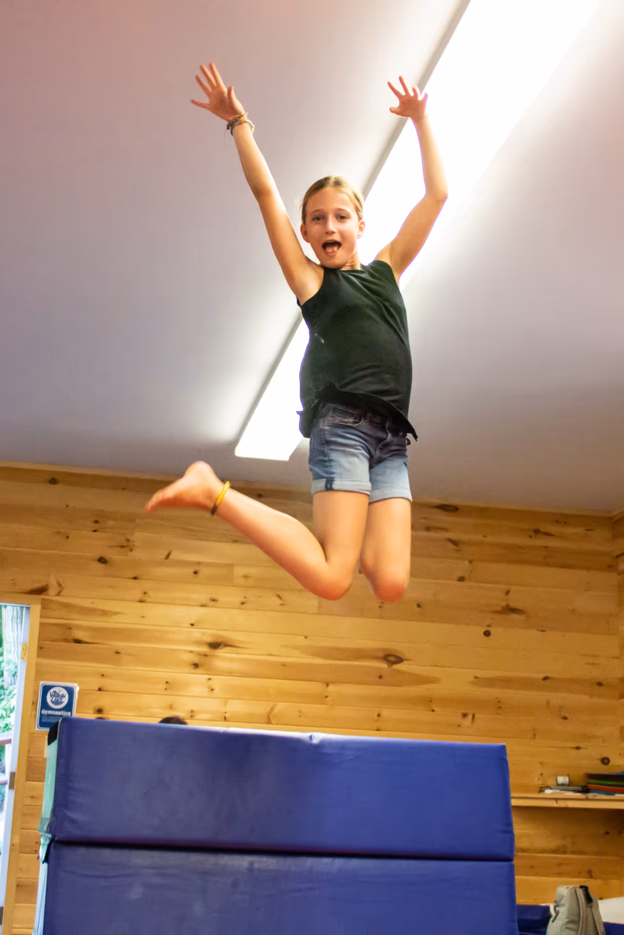 Young girl in a black tank top and denim shorts jumping in the air indoors over a blue gymnastics mat.