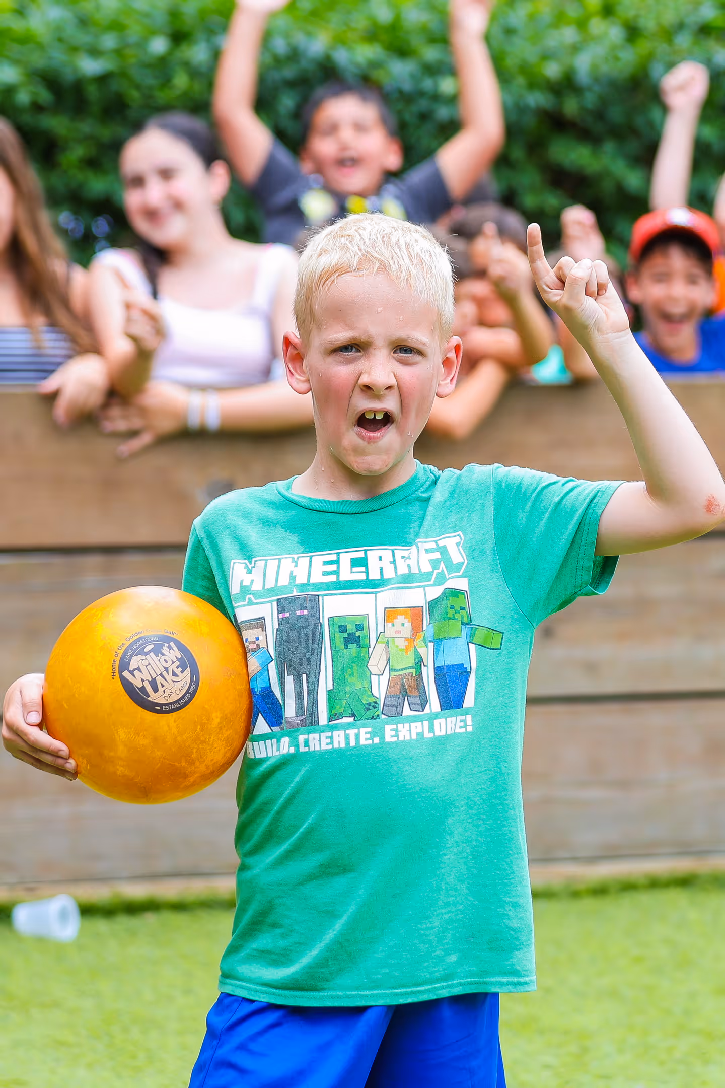 Blond boy in a green Minecraft shirt holding a yellow ball and raising one hand with a playful expression.