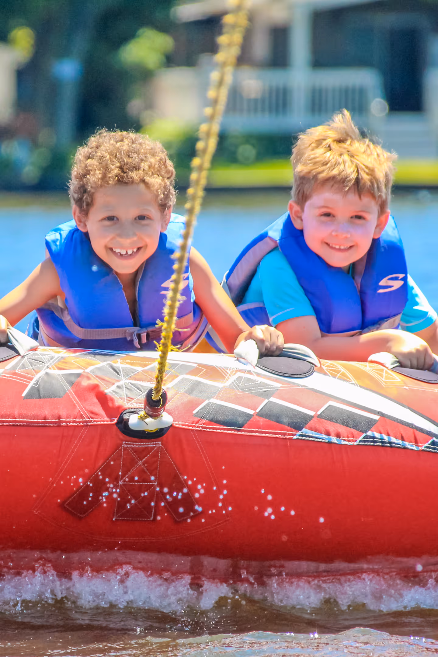 Two young boys wearing blue life jackets smiling while riding a red inflatable tube on water.