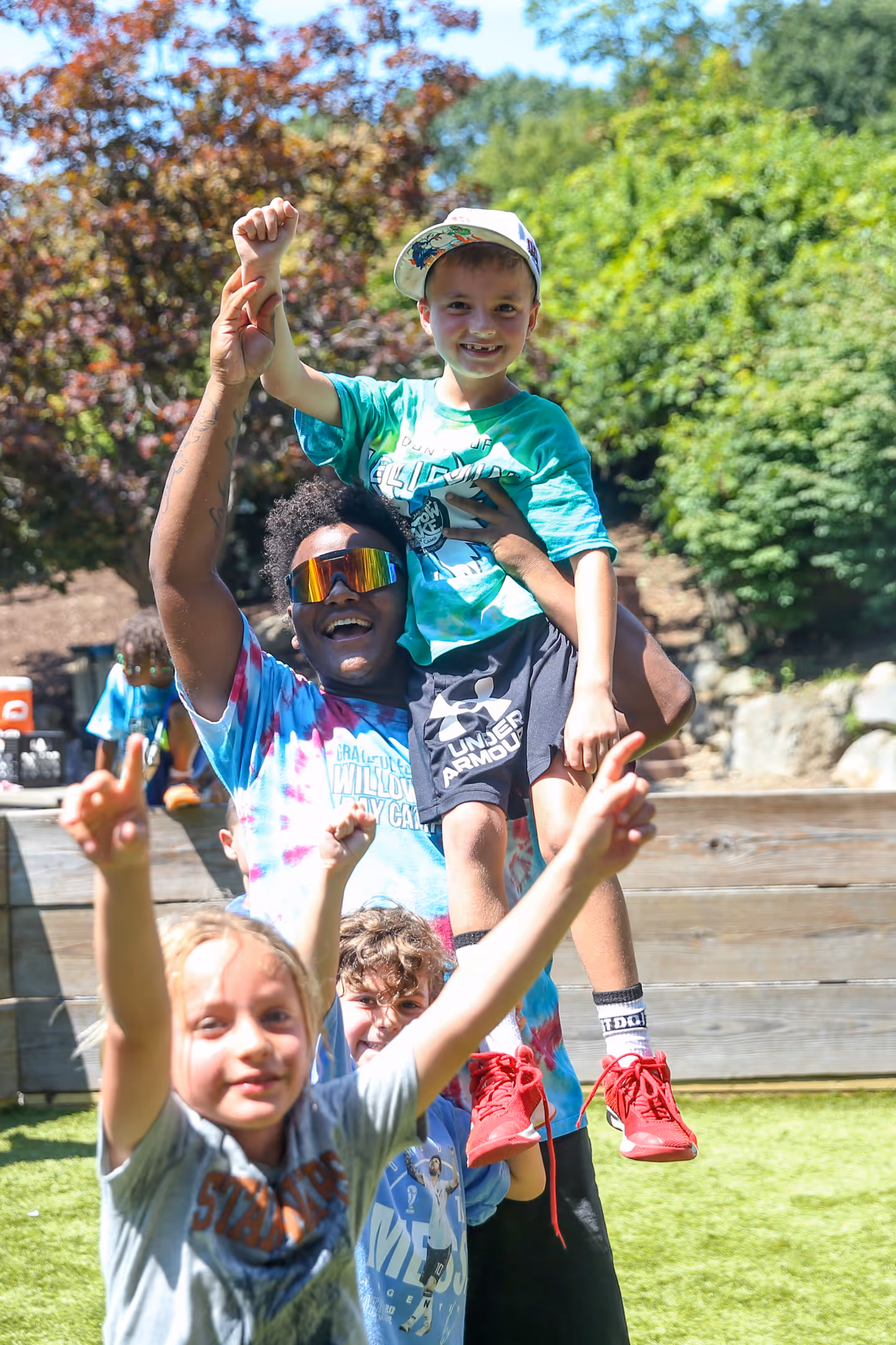 Camp counselor lifting a smiling boy wearing a cap with two children in front cheering outdoors.