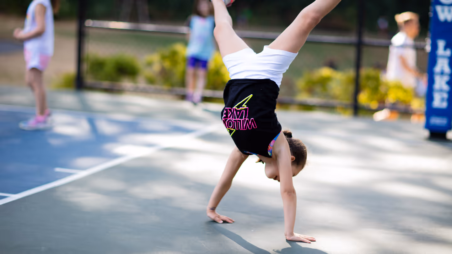 Girl doing a handstand on an outdoor basketball court with other children in the background.