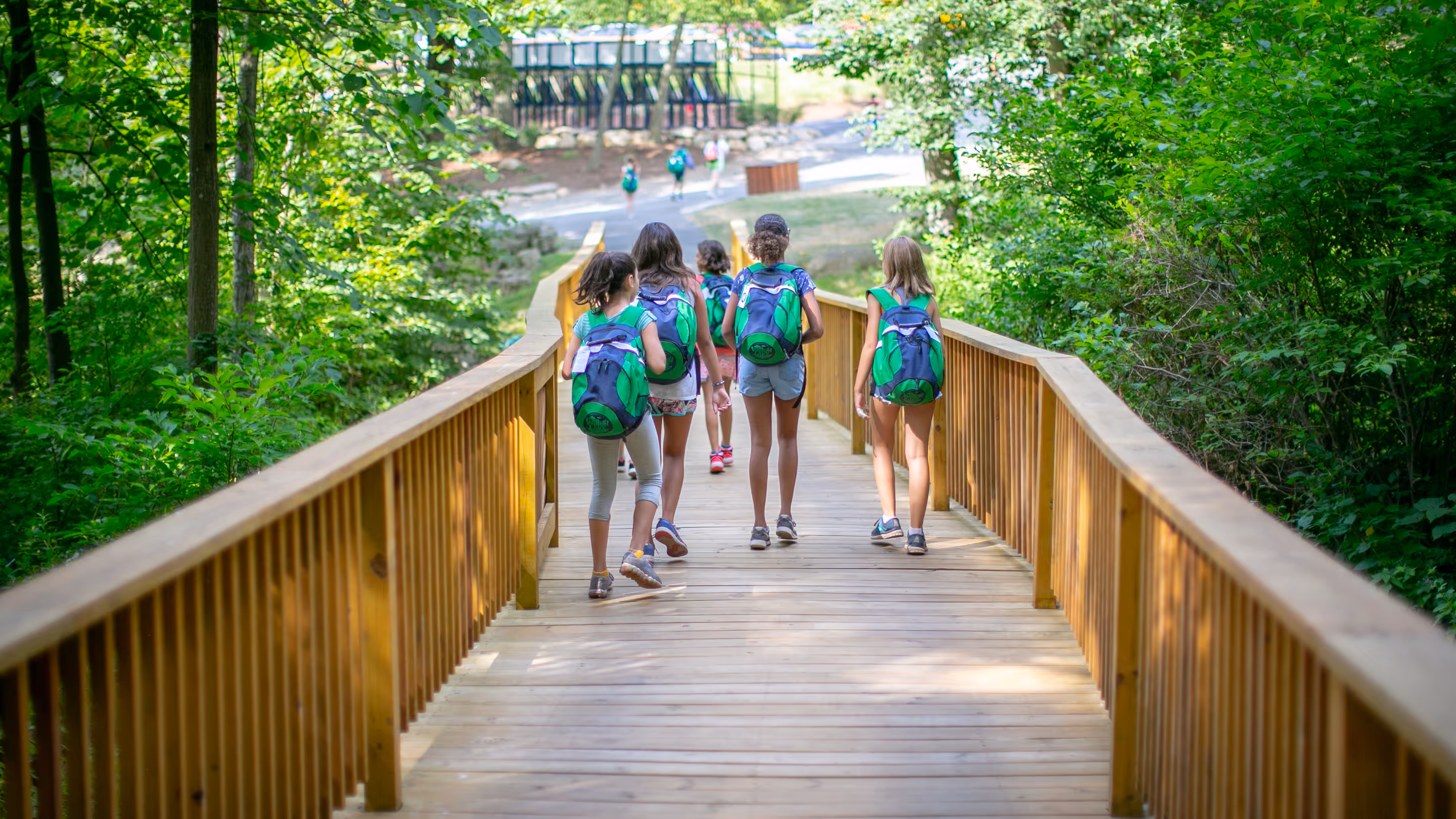 Group of children wearing matching green backpacks walking on a wooden bridge surrounded by green trees.