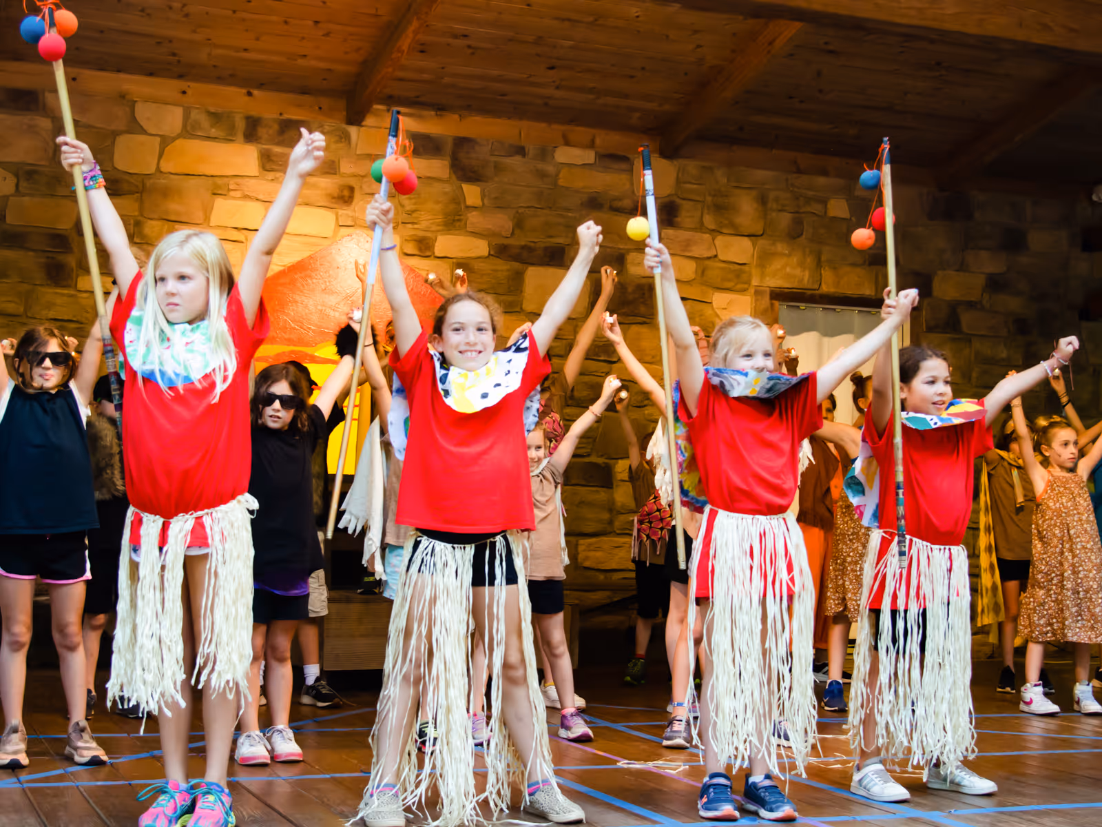 Group of children performing on stage, wearing red shirts, grass skirts, and holding decorated staffs with raised arms.