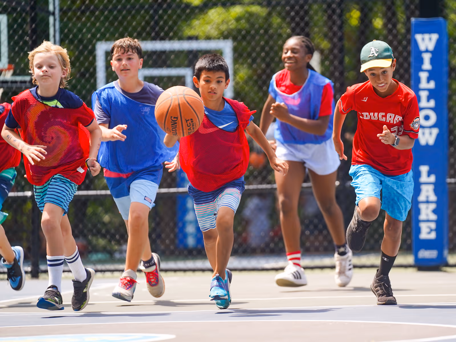 Five children wearing sports pinnies running on an outdoor basketball court with one boy dribbling a basketball.
