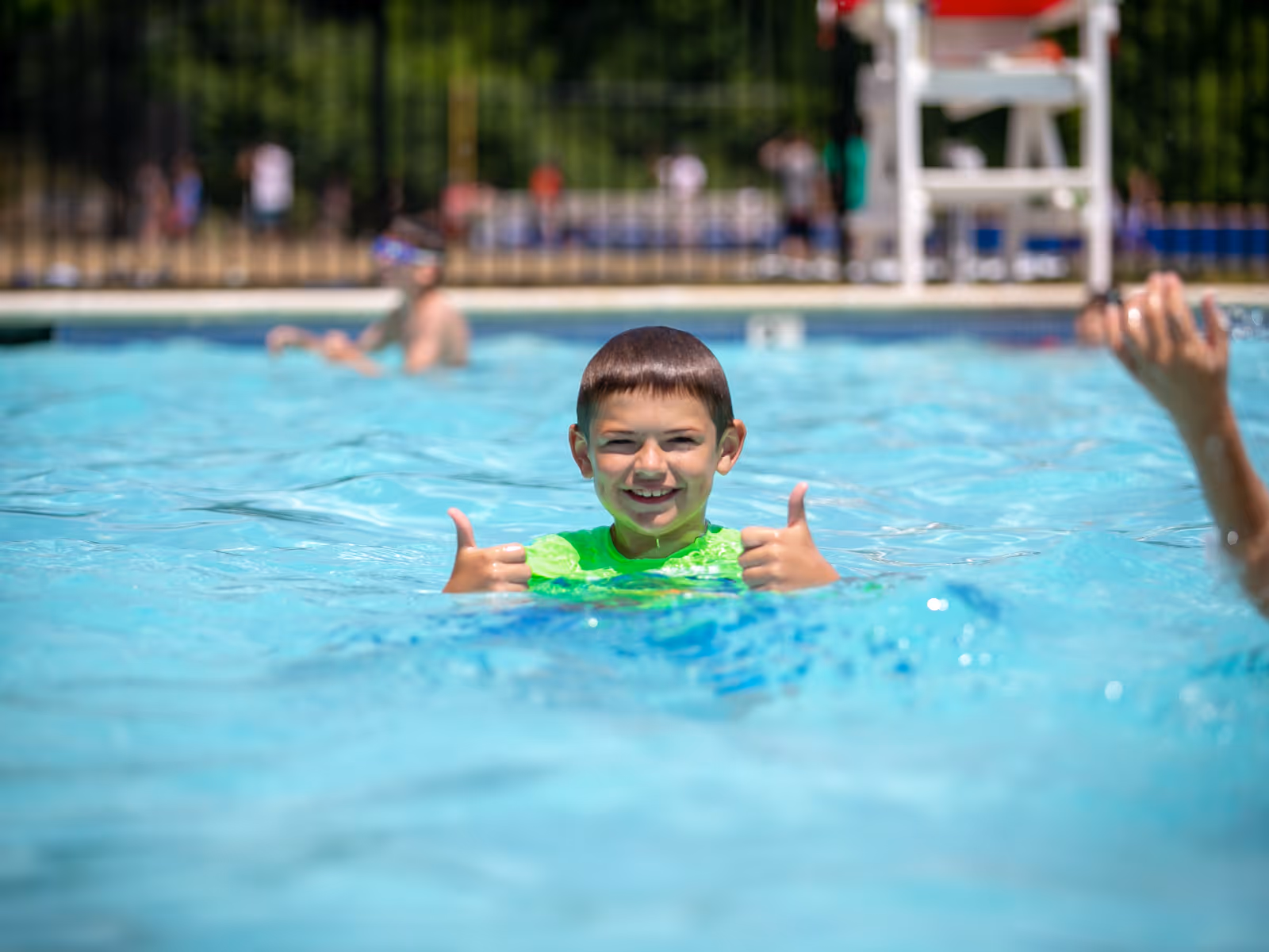 Smiling boy in a bright green shirt giving two thumbs up while swimming in a pool.