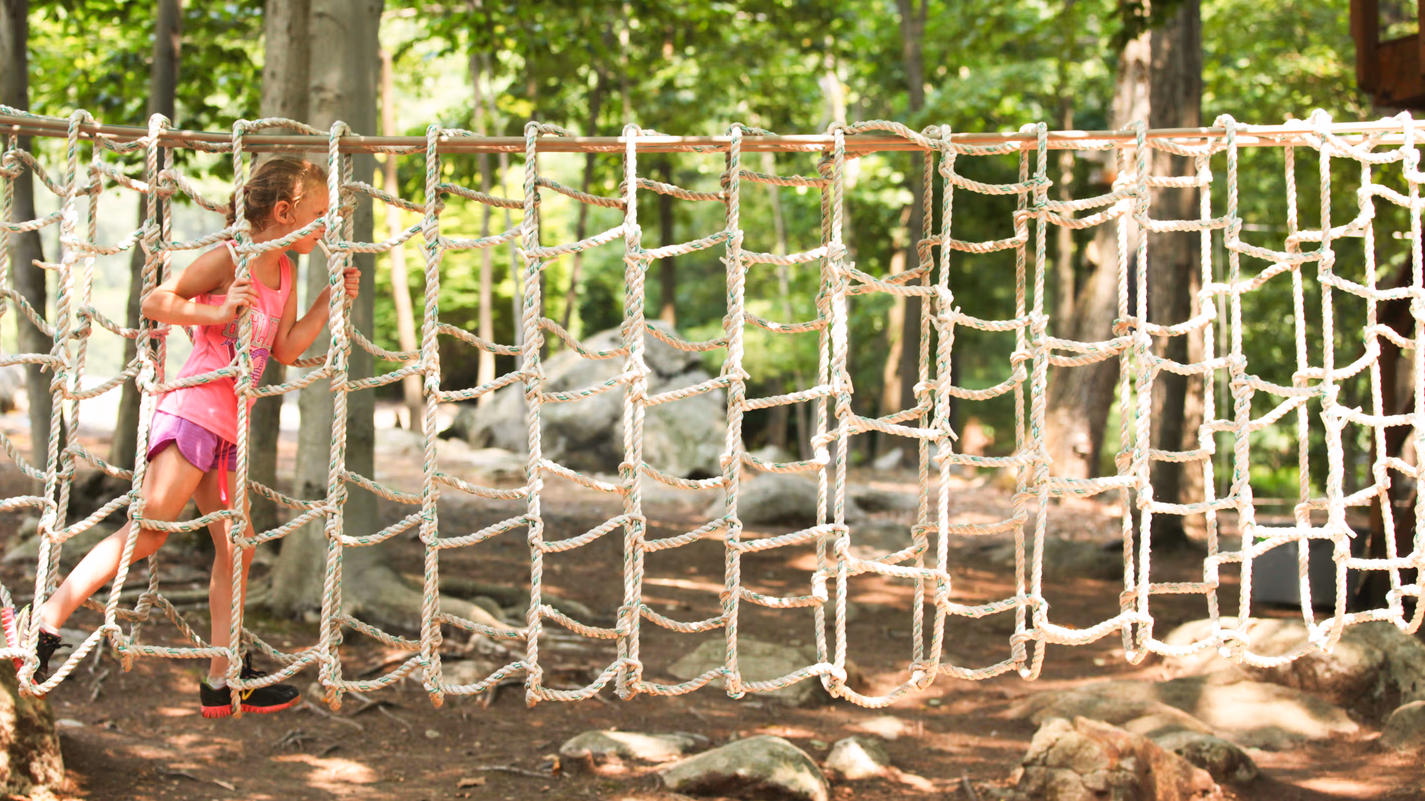 Young girl in pink shirt and shorts climbing on a rope net bridge in a wooded outdoor area.