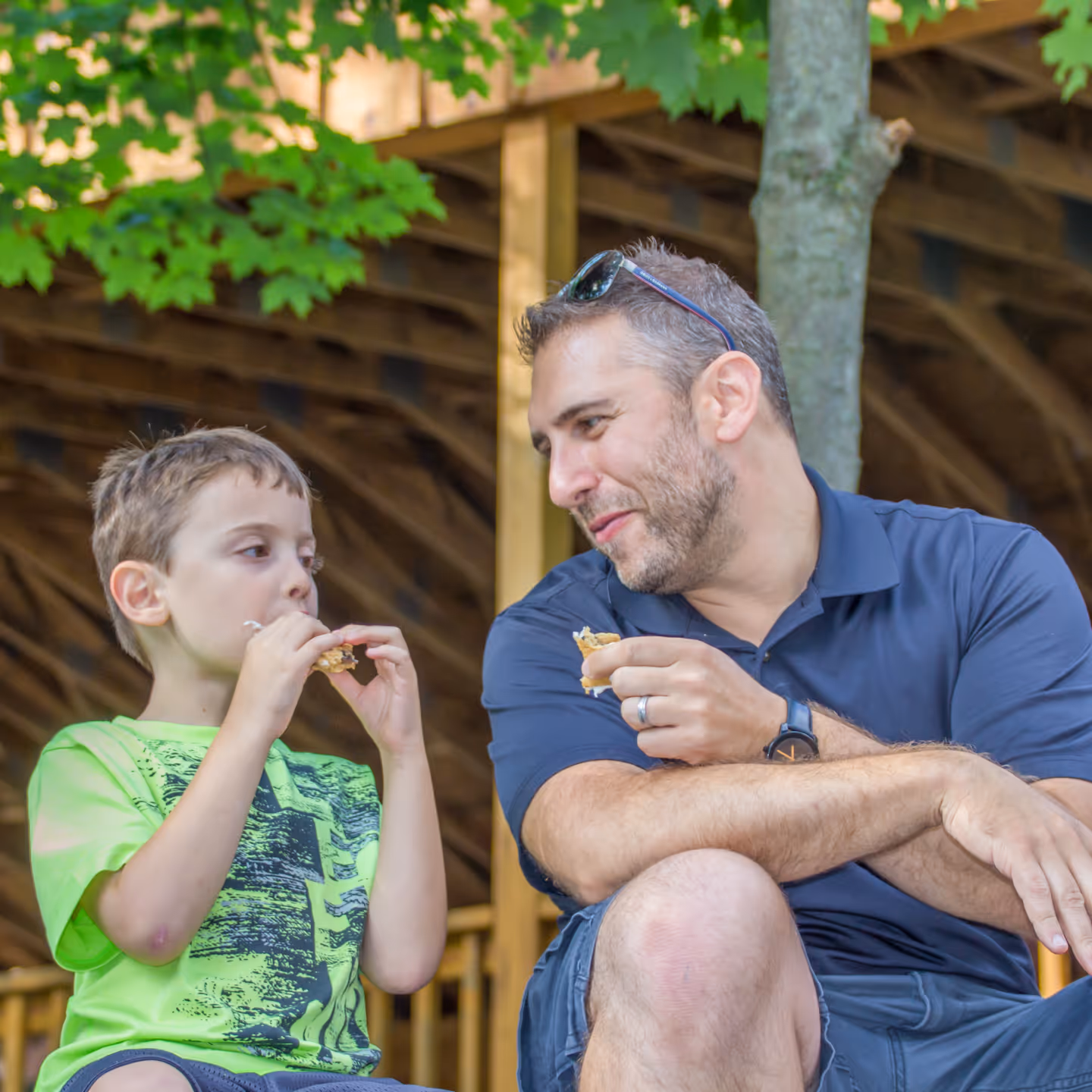 A man and a boy sitting outdoors by a tree eating s'mores together.
