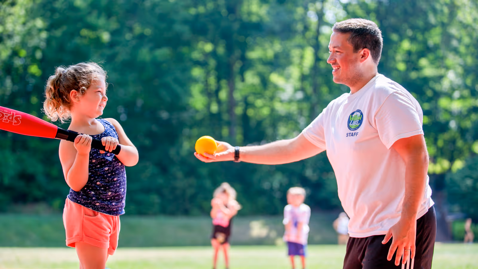 Camp staff member smiling and holding a yellow softball to a young girl preparing to bat outdoors.