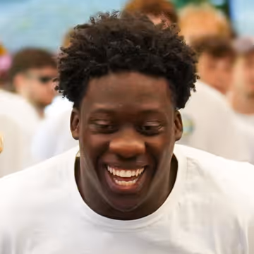 Young man with curly hair and a white shirt smiling broadly in an outdoor social setting.