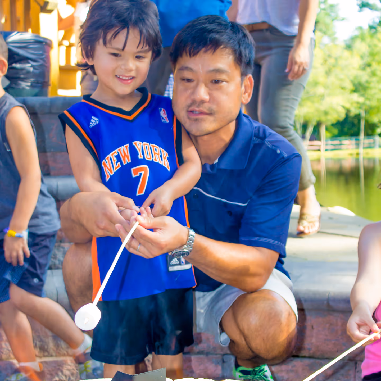 Father helping young boy roast a marshmallow outdoors near a pond.