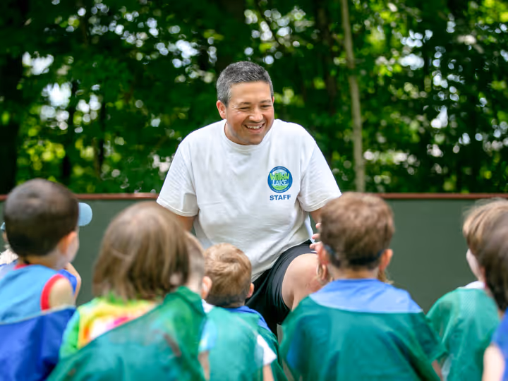 Smiling male camp staff member kneeling and engaging with a group of children outdoors.