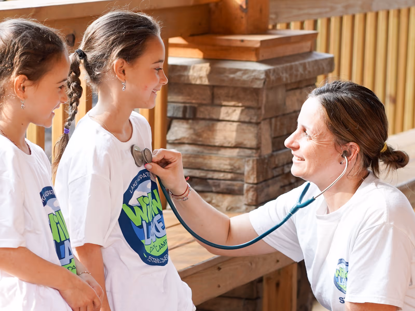 A nurse using a stethoscope to listen to a smiling girl's heartbeat while another girl watches, all wearing matching white camp T-shirts.