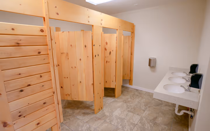 Clean restroom with light wood stall partitions and three sinks with soap dispensers on a tiled floor.