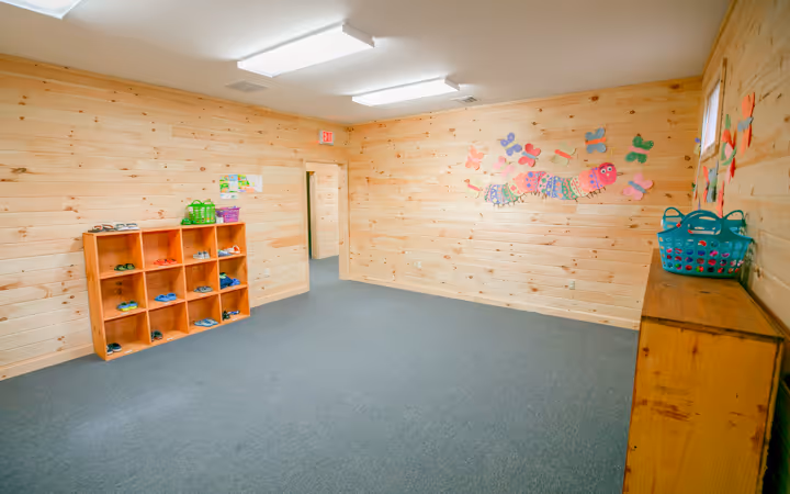 Bright room with wooden walls, blue carpet, cubbies with children's shoes, and colorful caterpillar and butterfly wall decorations.