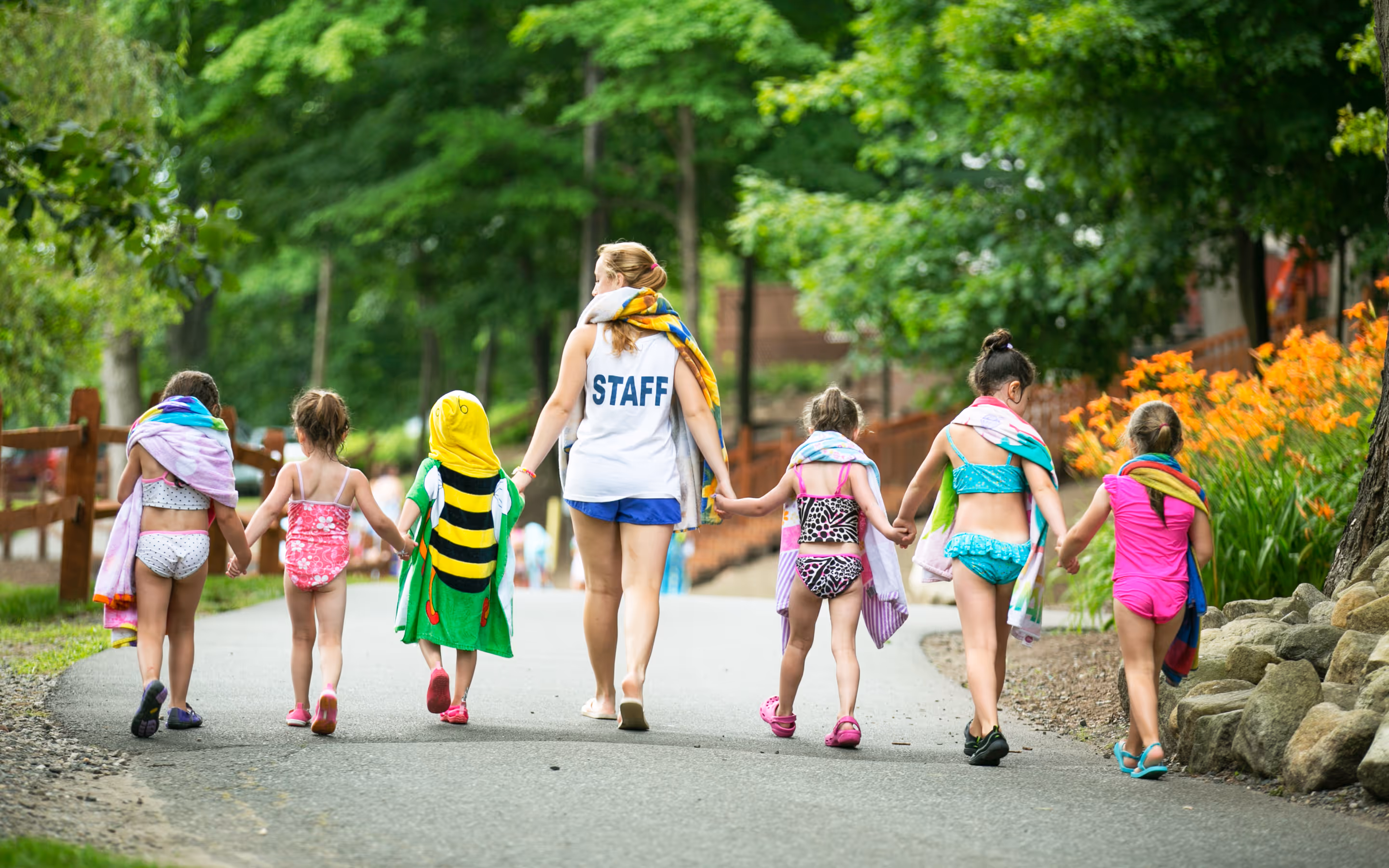 Staff member holding hands with six children in swimsuits and towels walking on a path surrounded by green trees and flowers.