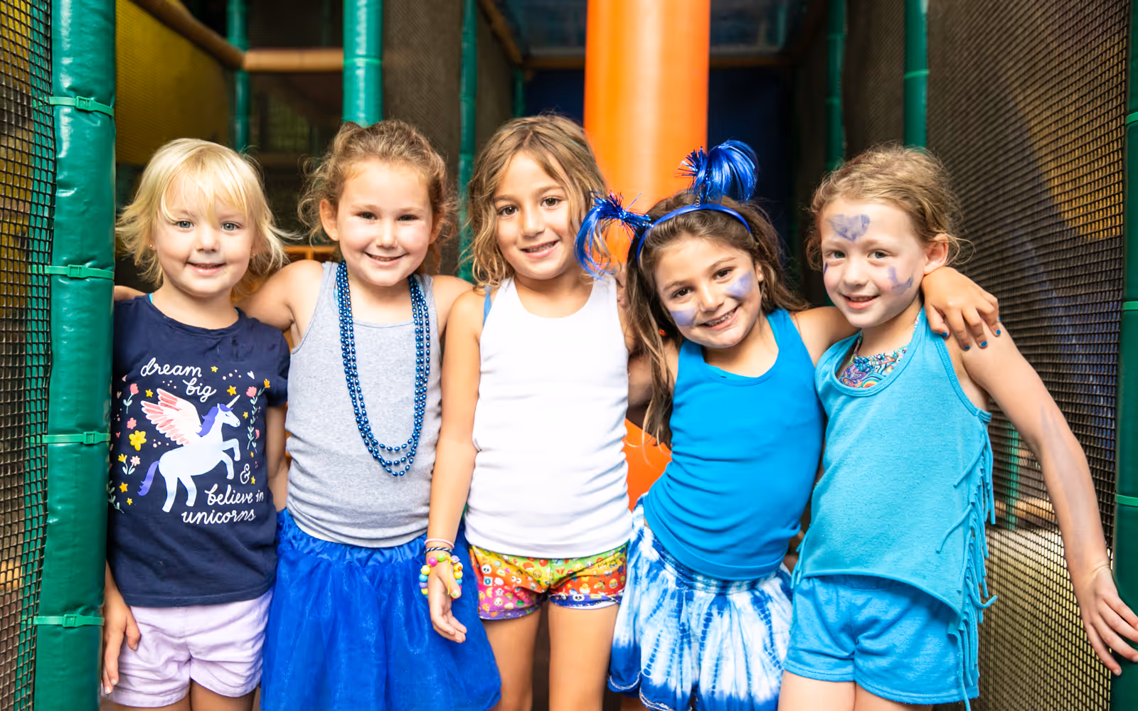 Five smiling young girls standing close together in colorful clothing and face paint inside a play area with green and orange padded poles.