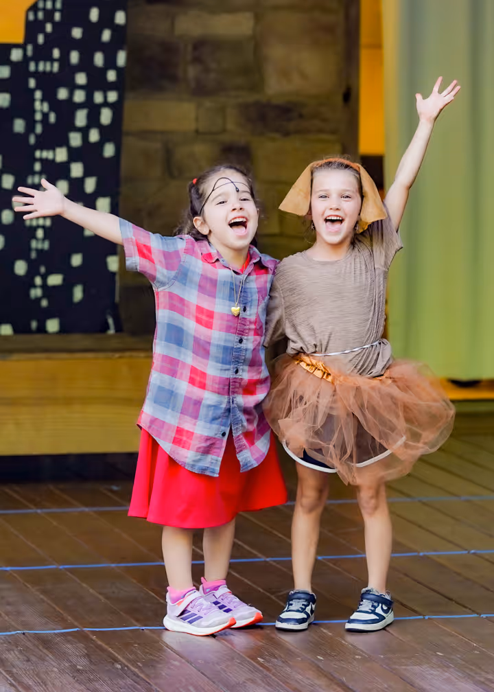 Two young girls on stage with arms raised, smiling and dressed in colorful costumes.
