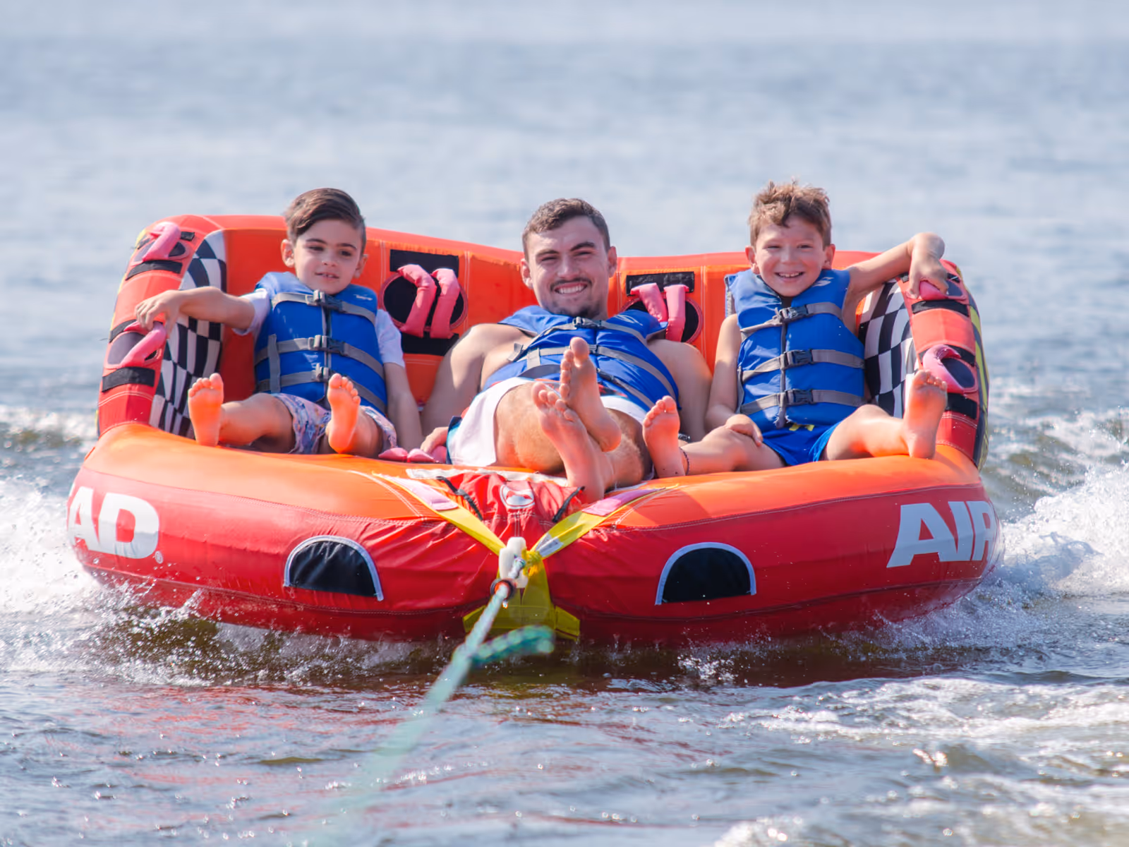 Two boys and a man wearing blue life jackets sitting on a red inflatable water tube being pulled on a lake.