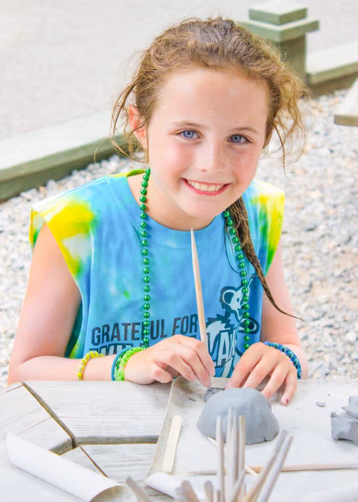 Young girl with blue eyes and a tie-dye shirt smiling while shaping clay at a table outdoors.