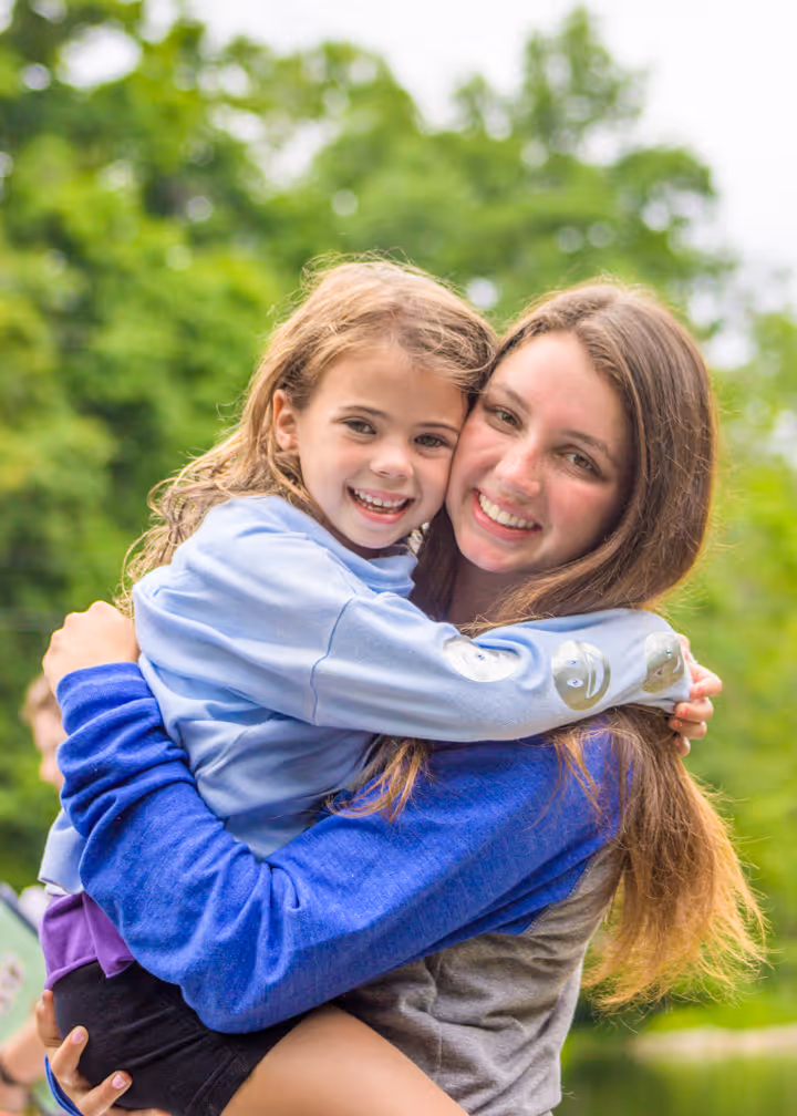 Young woman smiling while holding and hugging a smiling young girl outdoors with green trees in the background.