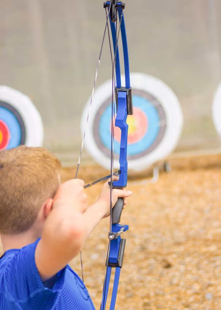 Boy aiming a blue recurve bow at an archery target during practice.