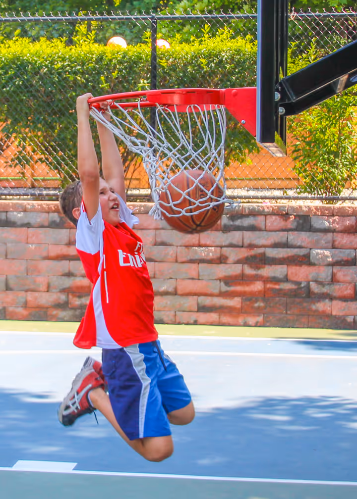 Boy in red and white shirt performing a slam dunk on an outdoor basketball court during the day.