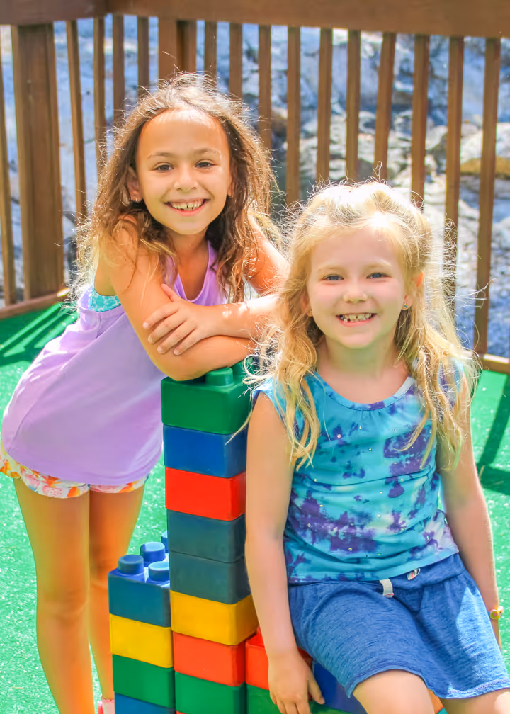 Two smiling young girls sitting and leaning on a colorful stacked block structure on green artificial grass with a wooden railing in the background.