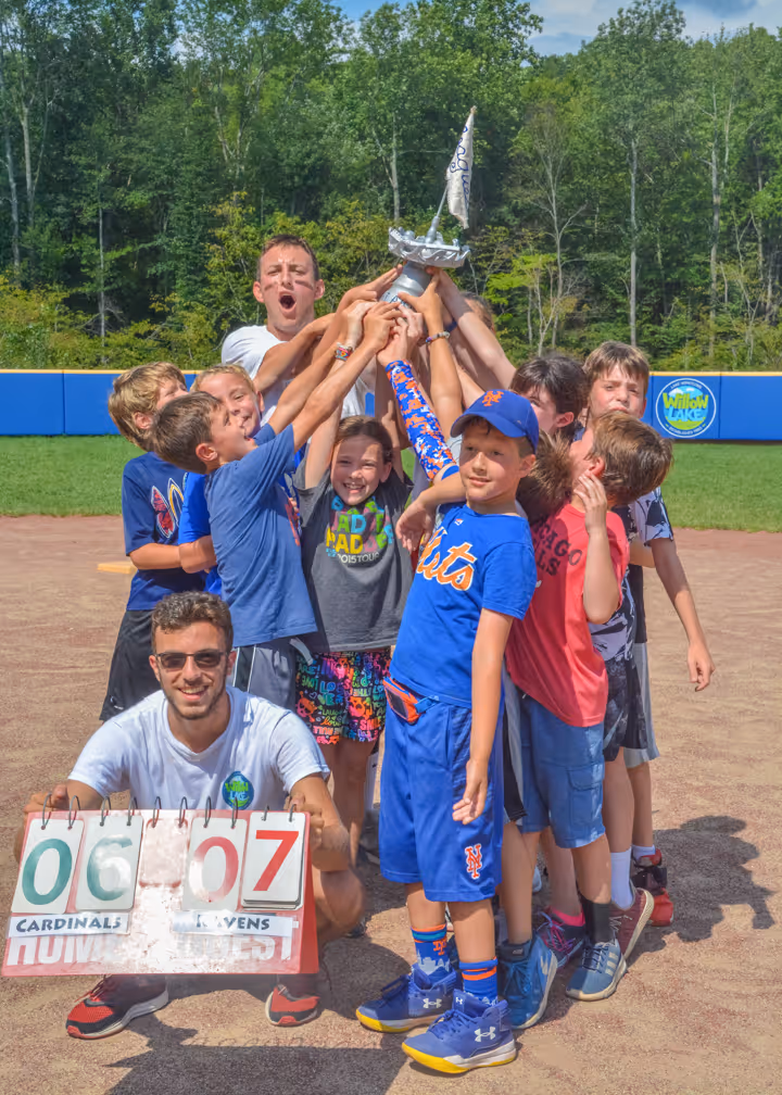Youth baseball team celebrating victory by raising a trophy together on the field with a scoreboard showing Ravens 7 and Cardinals 6.