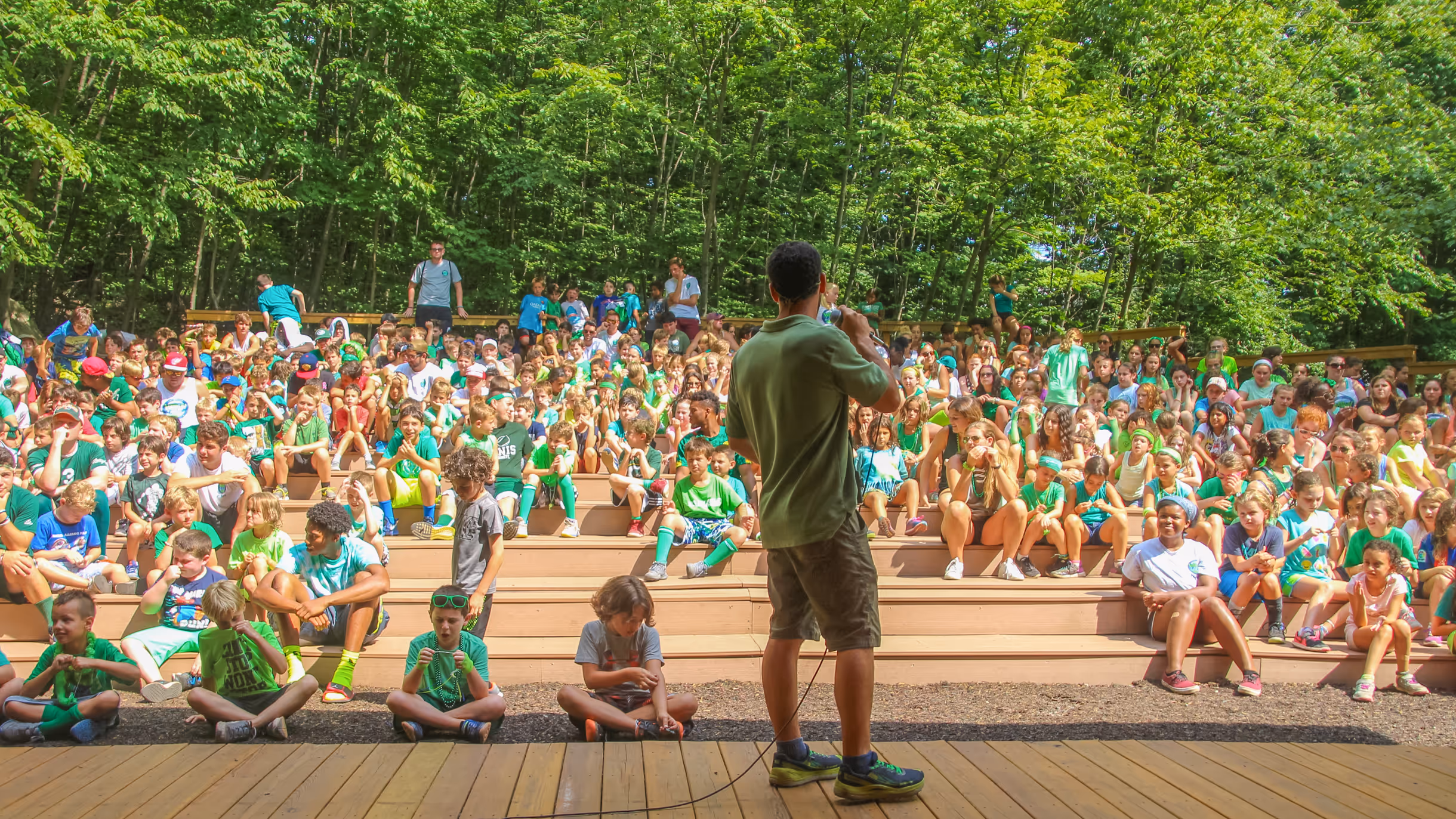 Man speaking with a microphone to a large outdoor audience of children seated on wooden bleachers surrounded by trees.