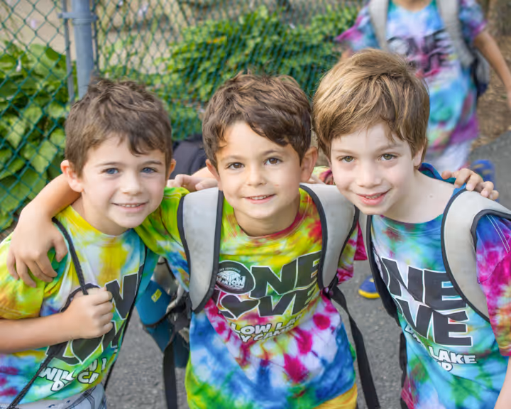 Three young boys smiling and wearing colorful tie-dye shirts with backpacks standing close together outdoors.