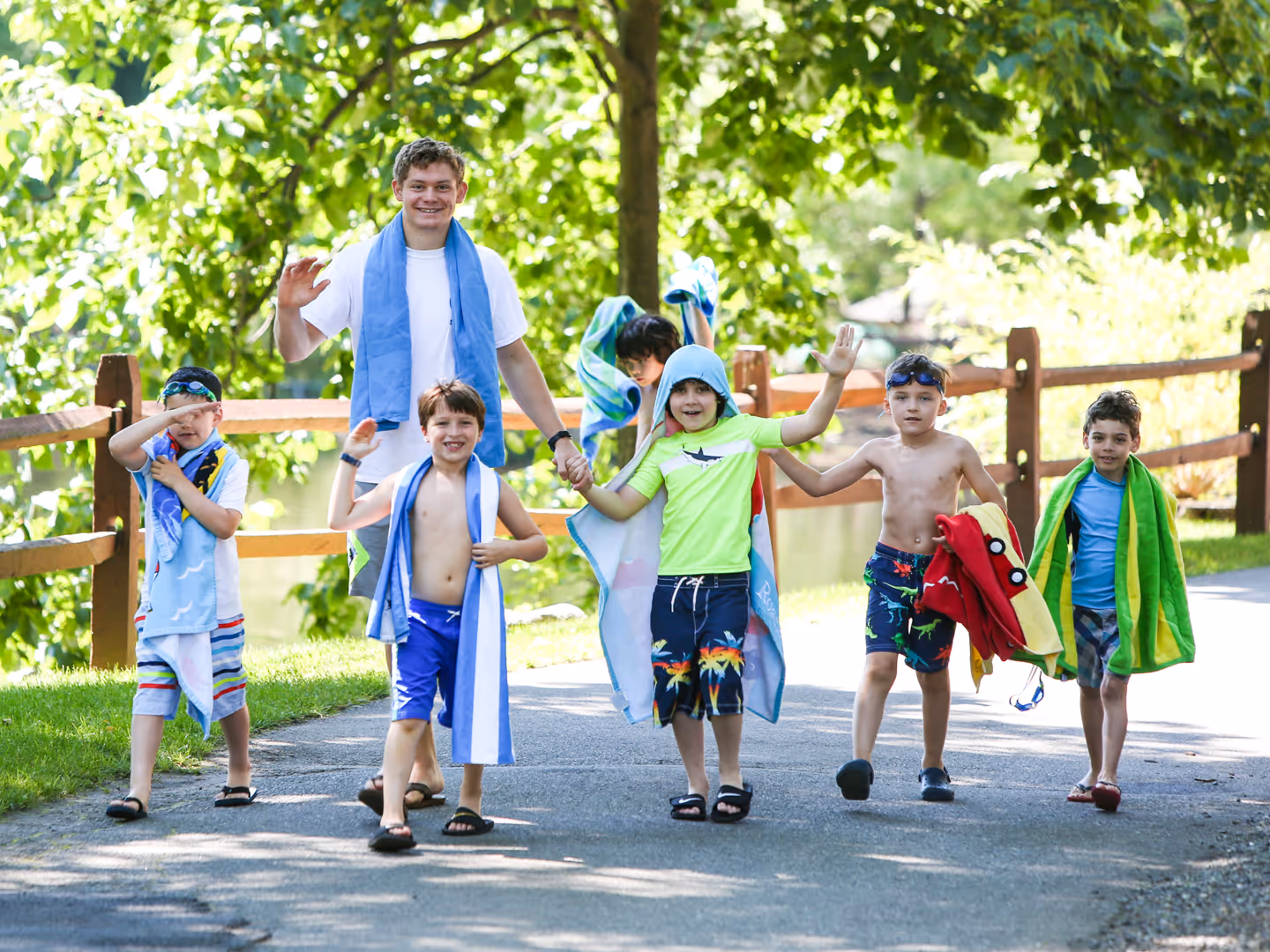 A group of six boys and one older boy walking on a paved path outdoors, wrapped in towels and swimwear, smiling and waving.