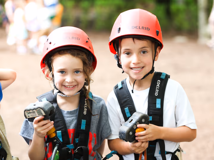 Smiling boy and girl wearing red helmets and harnesses, holding climbing equipment outdoors.