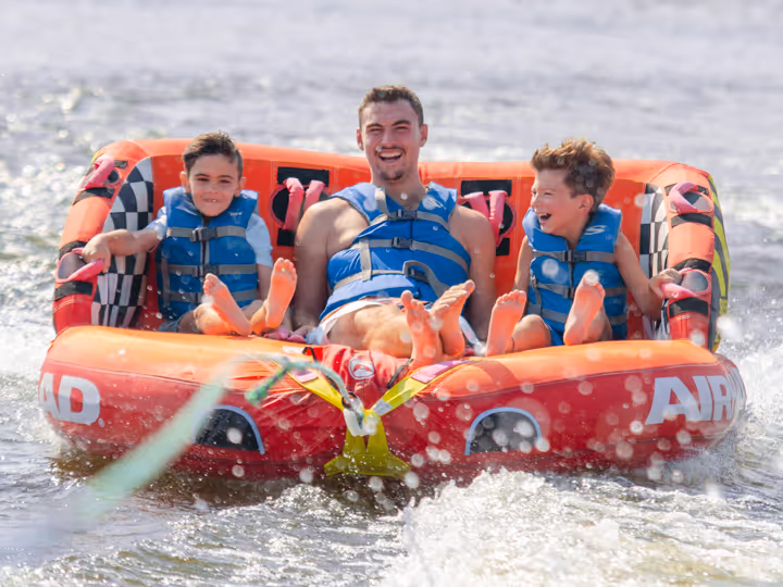 Adult male and two boys wearing life jackets, smiling and enjoying water tubing on an inflatable raft on a lake.