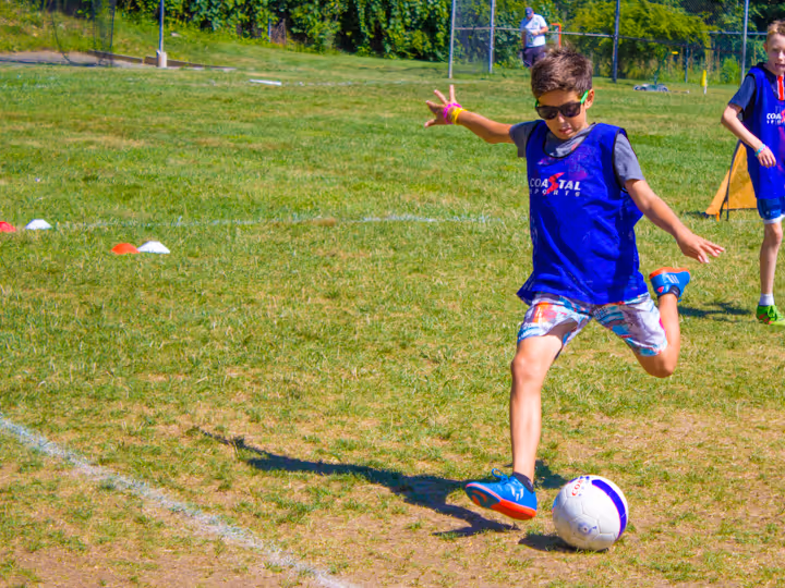 Boy wearing sunglasses and a blue sports vest kicking a soccer ball on a grassy field.