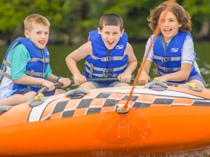 Three boys wearing blue life jackets riding an orange inflatable water tube on a lake.