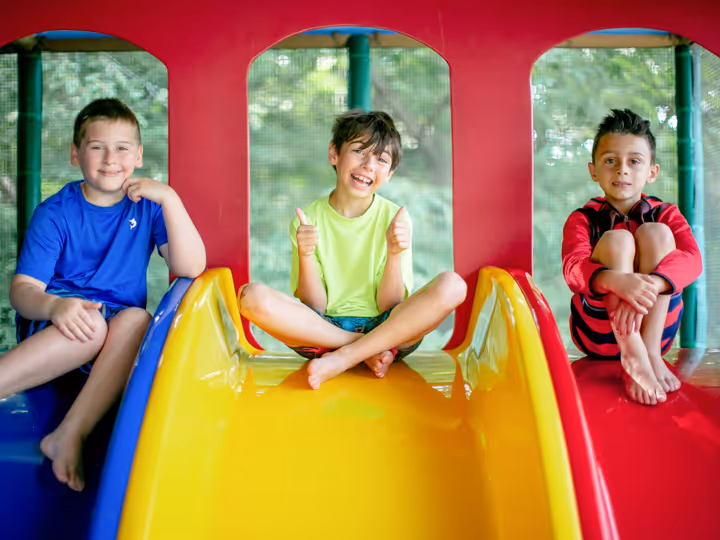 Three boys sitting on colorful playground slides, smiling and relaxed.