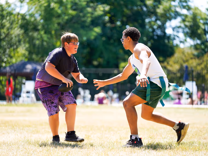 Two boys playing flag football outdoors, one holding the ball and the other reaching to grab a flag from his waist.