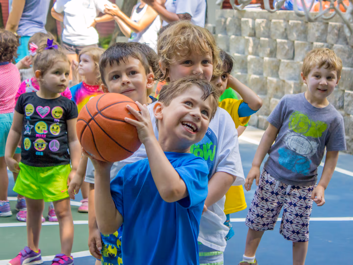 Group of young children playing basketball outdoors, with one boy smiling and holding the ball ready to shoot.