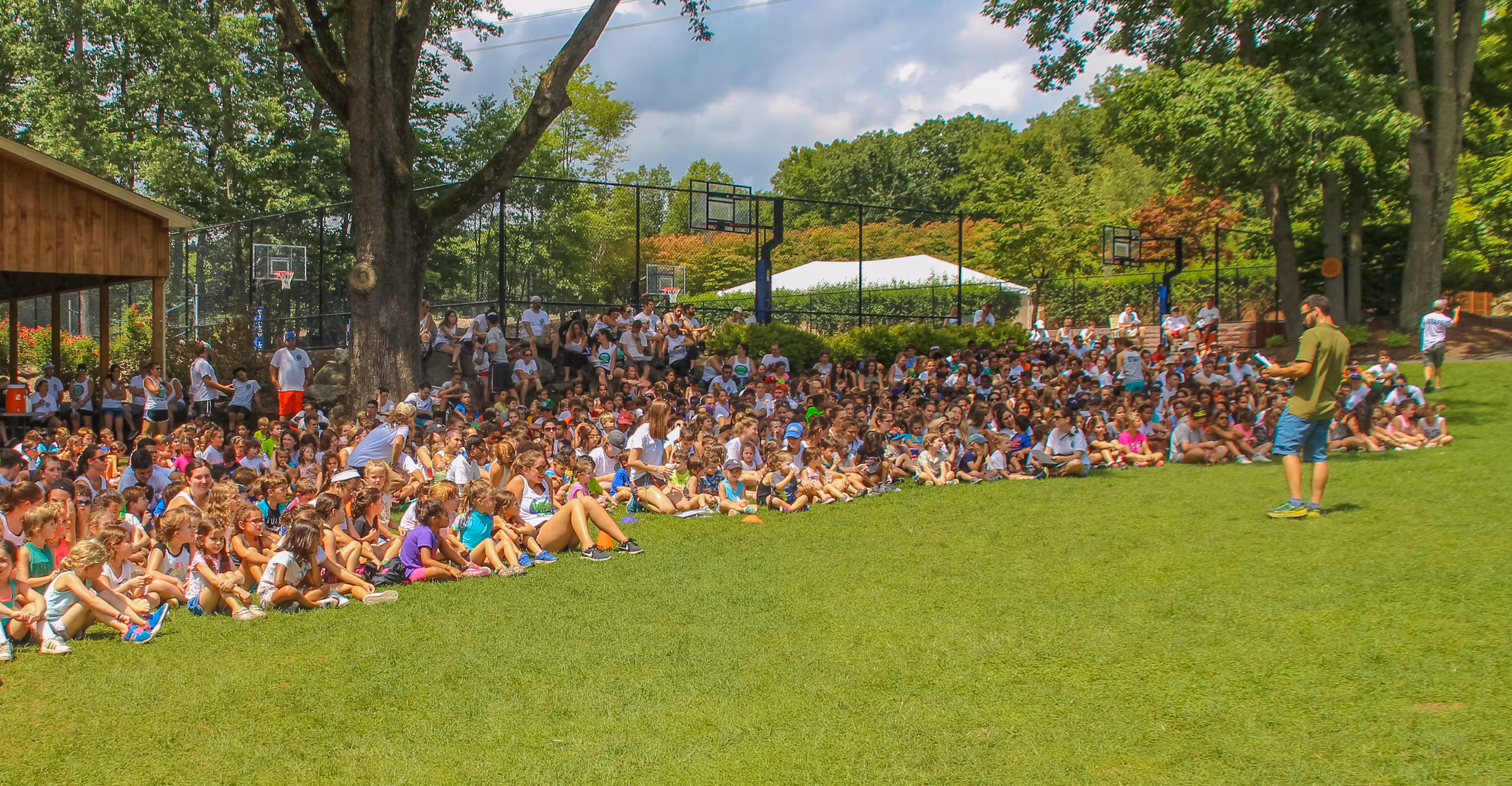 Large group of children and adults sitting on grass, listening to a standing man speaking at a camp assembly outdoors.