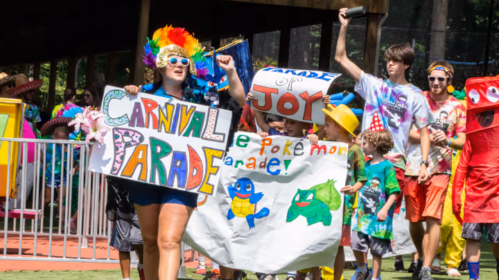 Group of people, including children and adults, participating in a colorful carnival parade with handmade signs and costumes, some featuring Pokémon characters.